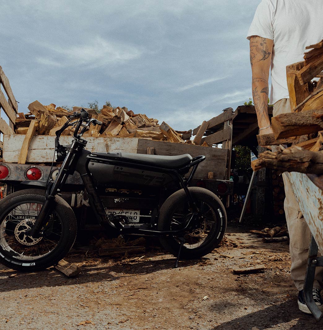 A SUPER73-B1G SE in Blackout sits parked behind a truck full of chopped wood logs. A man in light colored pants and a t-shirt stands to the right of the frame, facing the camera.