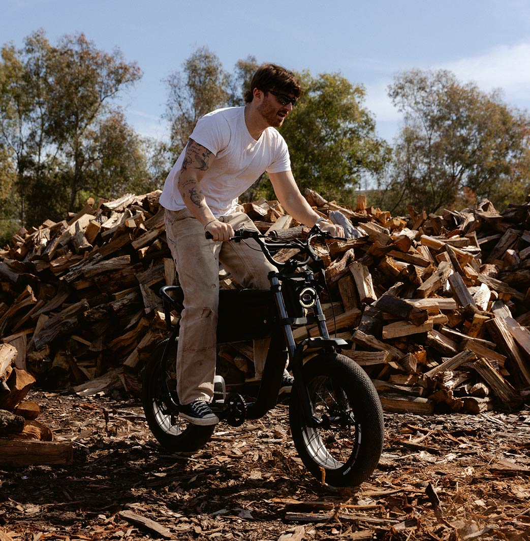 A man in light colored pants and a t-shirt stands while riding a SUPER73-B1G SE in Blackout. A large pile of wooden logs is visible in the background.