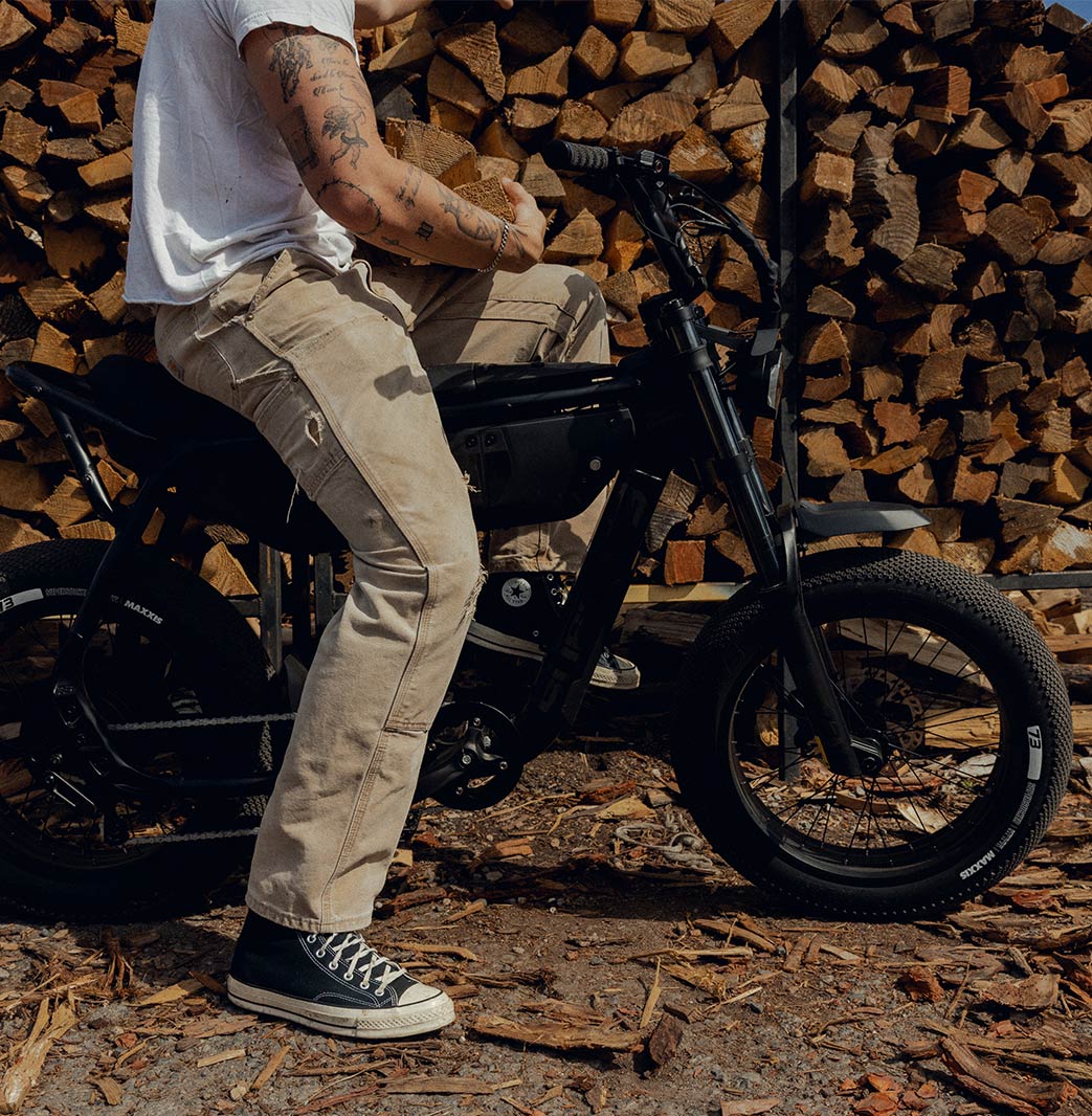 A man in light colored pants and a t-shirt sits on a SUPER73-B1G SE in front of a large pile of wooden logs.