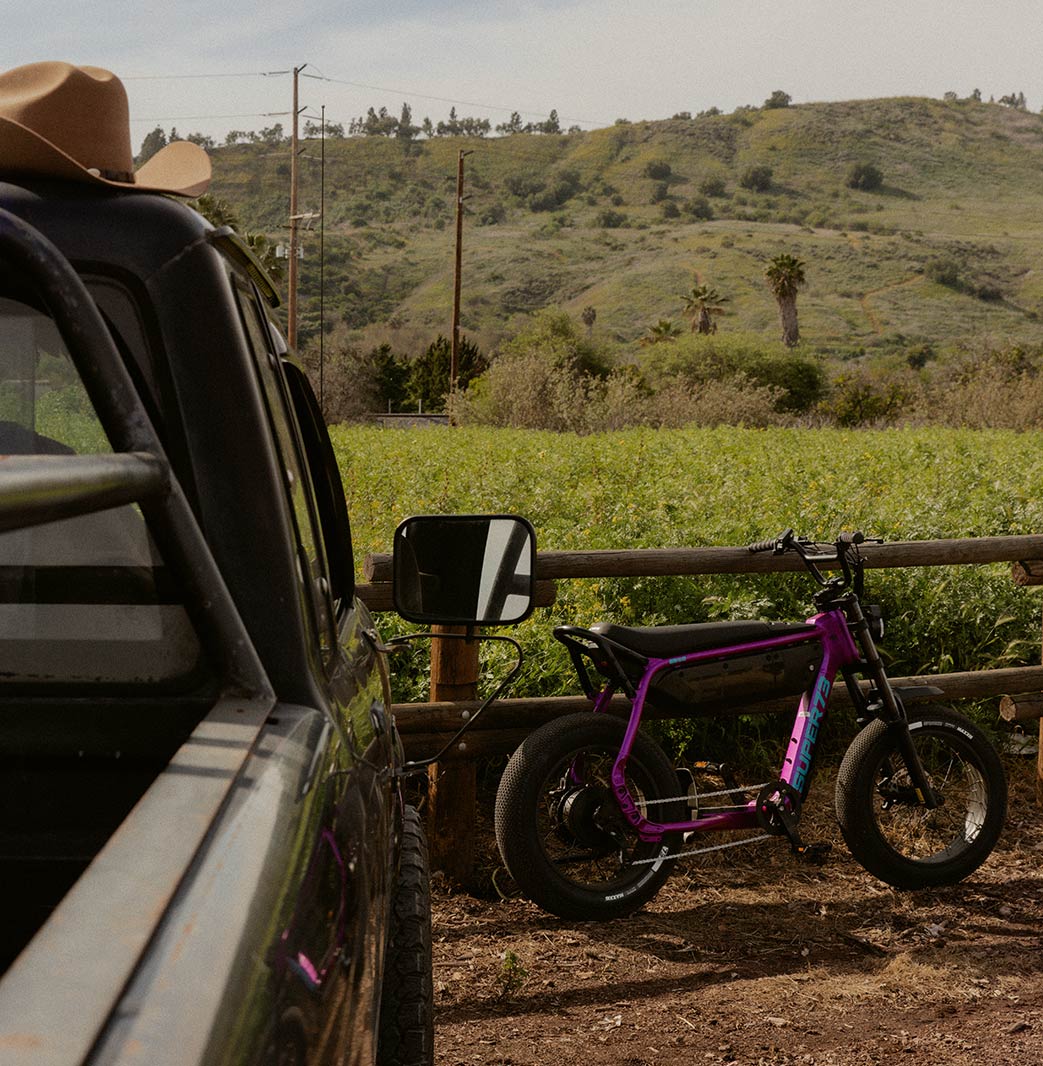 A black pickup truck is parked on the left-hand side of the frame with a brown cowboy hat sitting atop it. To the right, a SUPER73-B1G SE in Cullenberry is parked against a wooden fence with a large green field in the background.