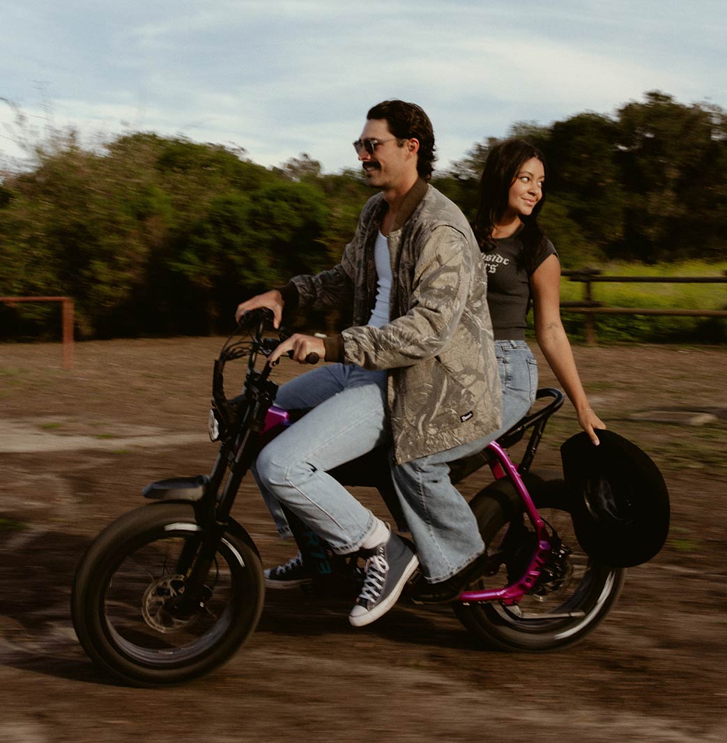A man wearing jeans, a light colored top and a light colored camo print jacket rides a SUPER73-B1G-SE in Cullenberry on a dirt path. A woman wearing jeans and a black t-shirt sits on the back of the bike, smiling and holding a black cowboy hat.
