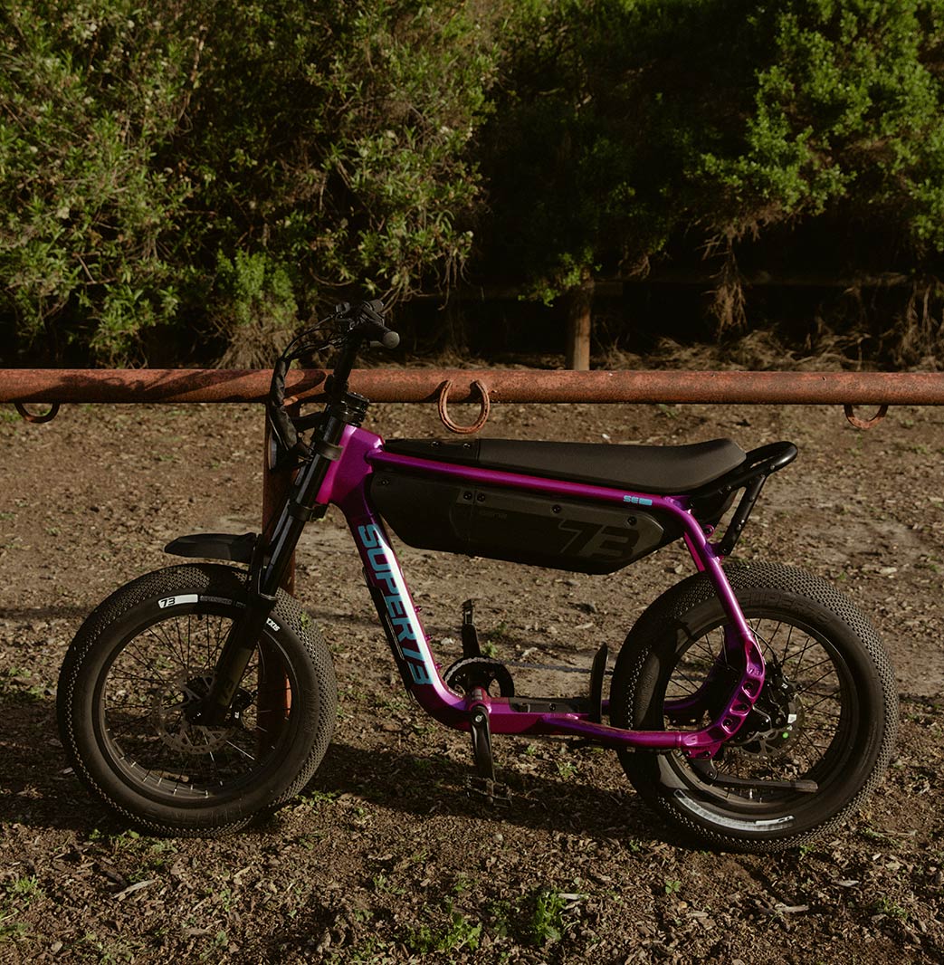 A SUPER73-B1G SE in Cullenberry sits on a dirt path leaning against a metal fence with a horseshoe on it.