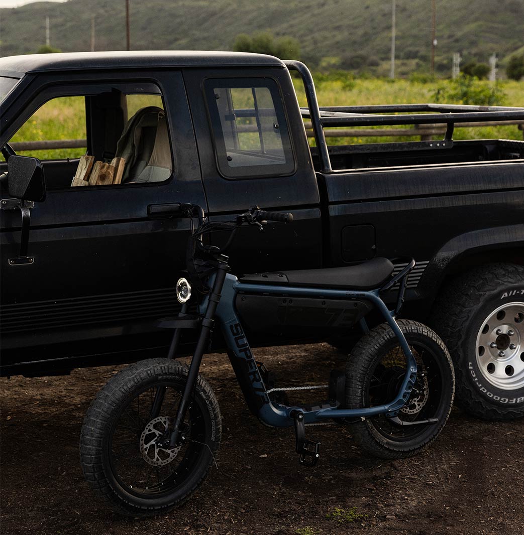 A black pickup truck is parked along a dirt path with a green field in the background. A SUPER73-B1G leans against the truck.