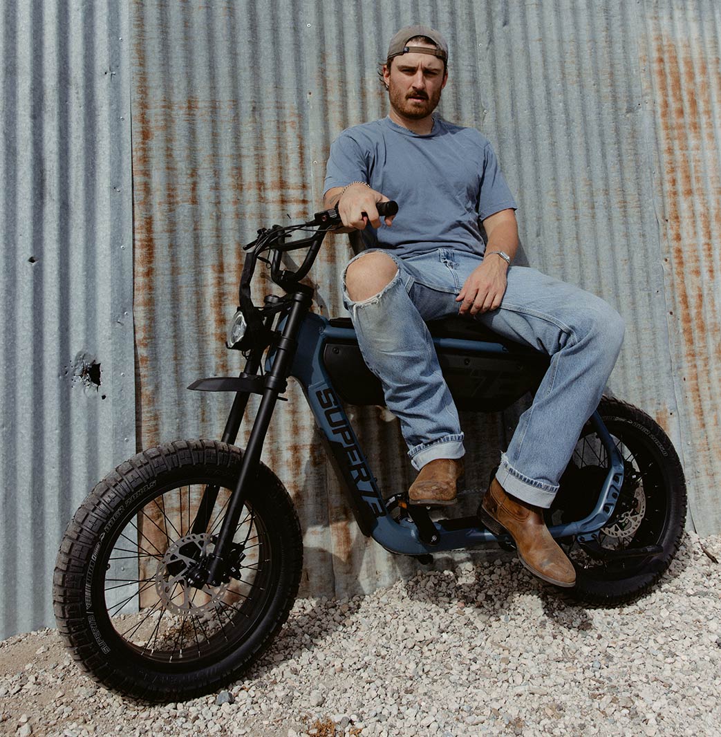 A man in light colored jeans and a t-shirt sits sideways on a SUPER73-BIG leaning against a metal wall on a gravel path.