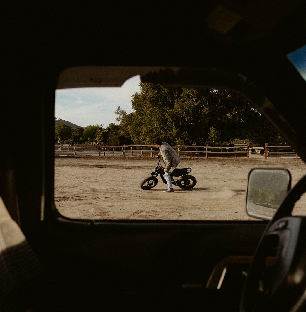 A man in light colored jeans and a jacket riding a SUPER73-B1G in an open area on dirt, photographed from inside the cab of a truck.
