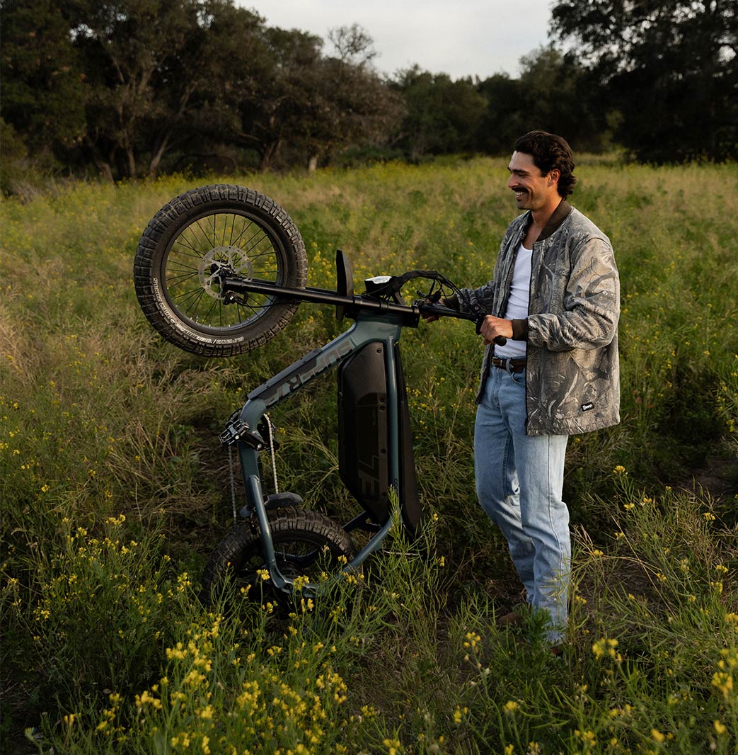 A man wearing light colored jeans, a white top and a light colored jacket stands in a green field while holding the handlebars of a SUPER73-M1D. The ebike is vertical and resting only on its back tire with the front tire facing up.