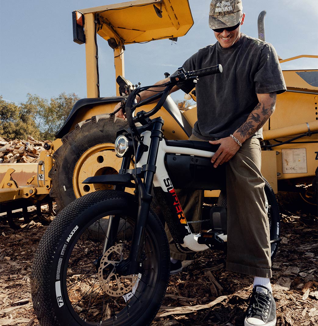 A man in green pants and a dark t-shirt is sitting on the SUPER73-M1D SE ebike in Baja with a large yellow piece of machinery parked behind him.