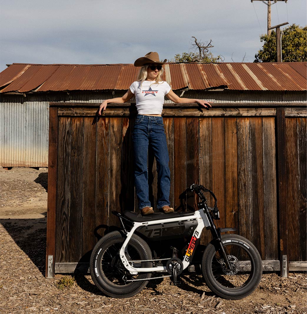 A woman wearing jeans, a white t-shirt, and a brown cowboy hat is standing on the seat of a SUPER73-MZFT SE ebike. The bike is parked next to a wooden fence, which the woman is leaning against.