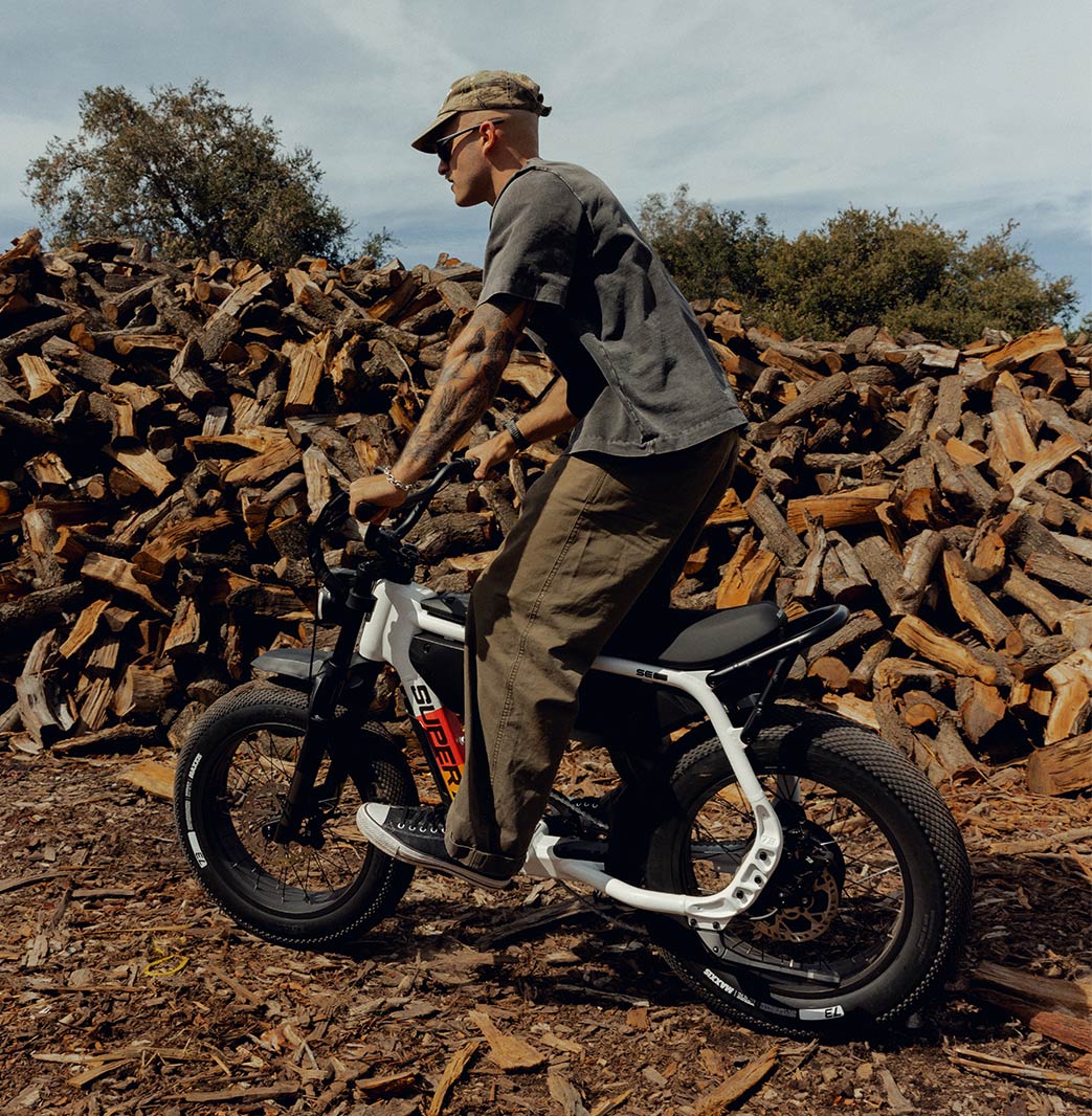 A man wearing green pants and a dark t-shirt riding a SUPER73-MZFT SE ebike. The man is standing on the pedals and a stack of wood logs is visible in the background.