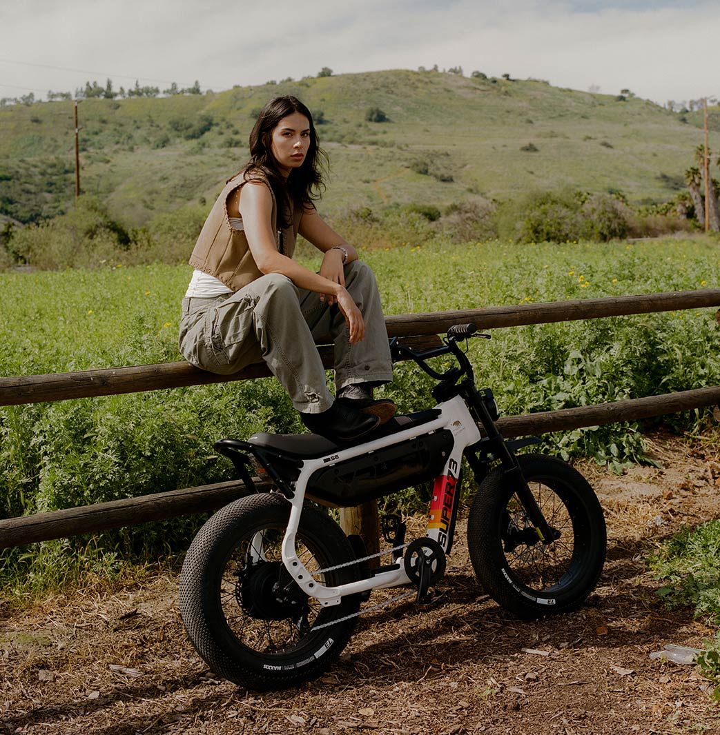 A woman wearing light green pants and a light colored top is sitting on a wooden fence with her feet resting on the seat of a SUPER73-MZFT SE ebike. The bike is parked in front of the fence and a green field and hills are visible in the background.