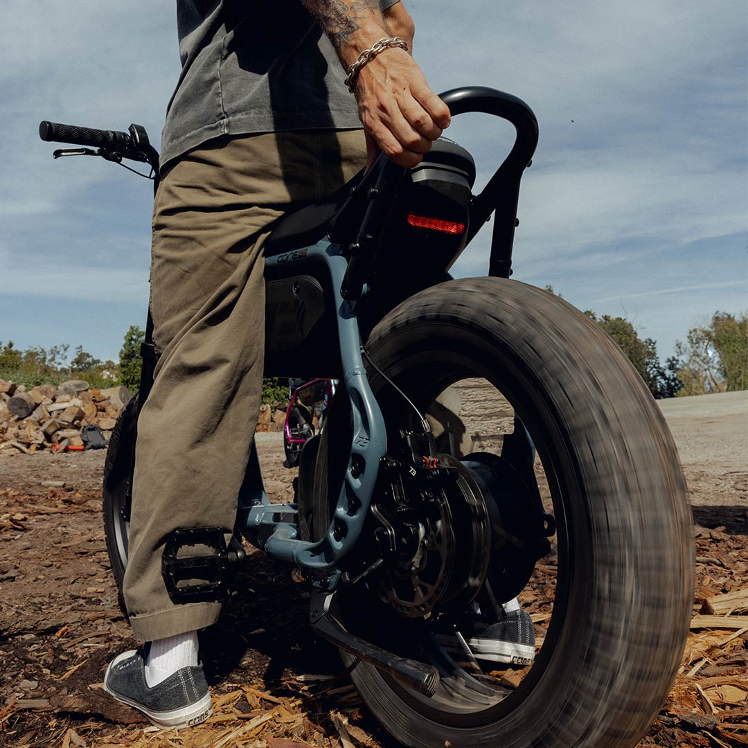 A man wearing dark green pants and a dark colored t-shirt is straddling a SUPER73-M1D ebike with his hand on the rear of the frame. The back tire appears to be spinning.
