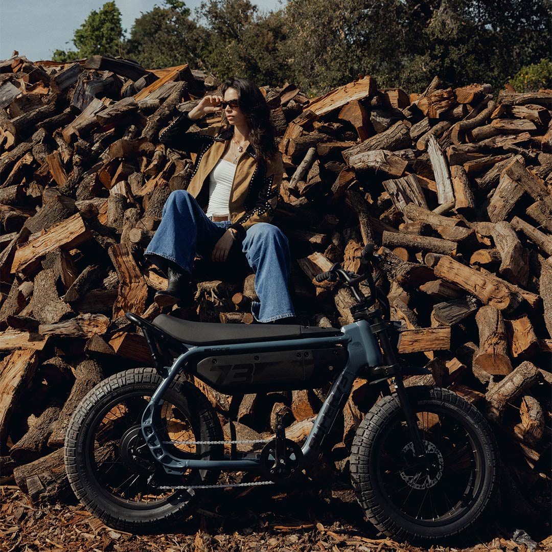 A woman wearing jeans and light colored top and jacket is sitting on a large pile of wooden logs. A SUPER73-M1D ebike is parked in front of her at ground level.
