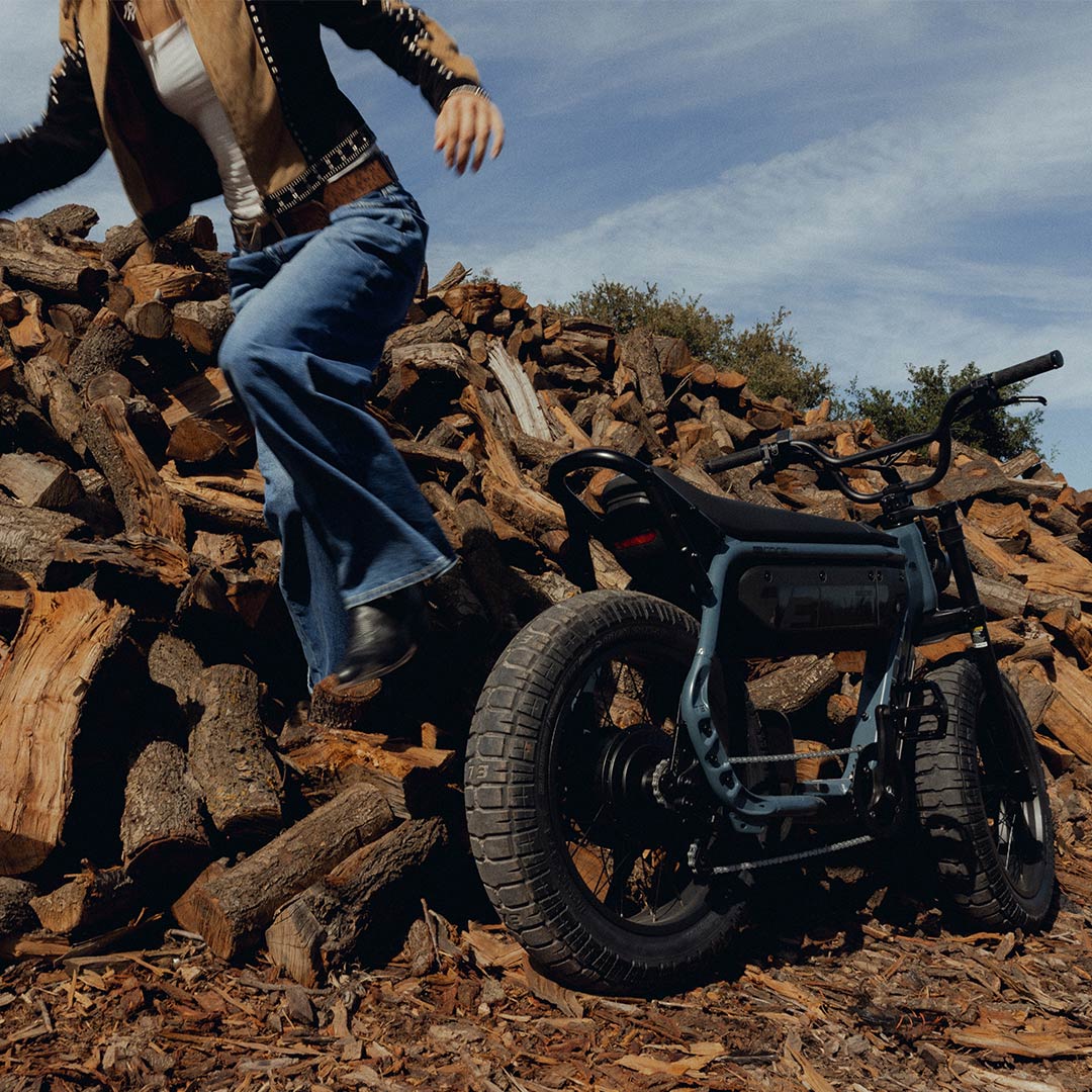A woman wearing jeans and light colored top and jacket is jumping down from a large pile of wooden logs. A SUPER73-M1D ebike is parked in front of the pile on the right-hand side of the frame, facing away from the camera.