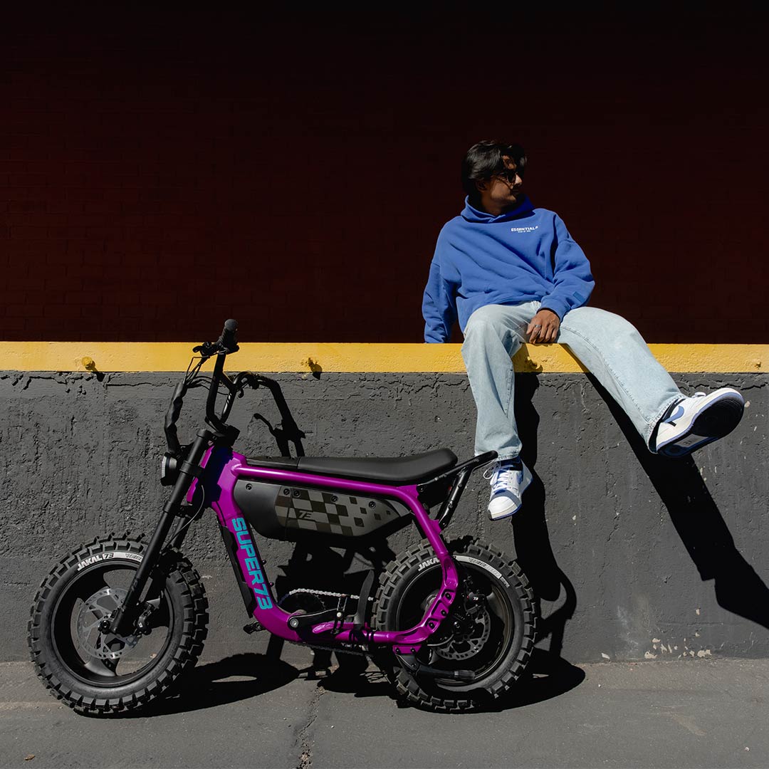 A man in jeans and a blue sweatshirt sits on a concrete wall with a yellow stripe painted across it. A SUPER73-MZFT ebike is parked in front of him.