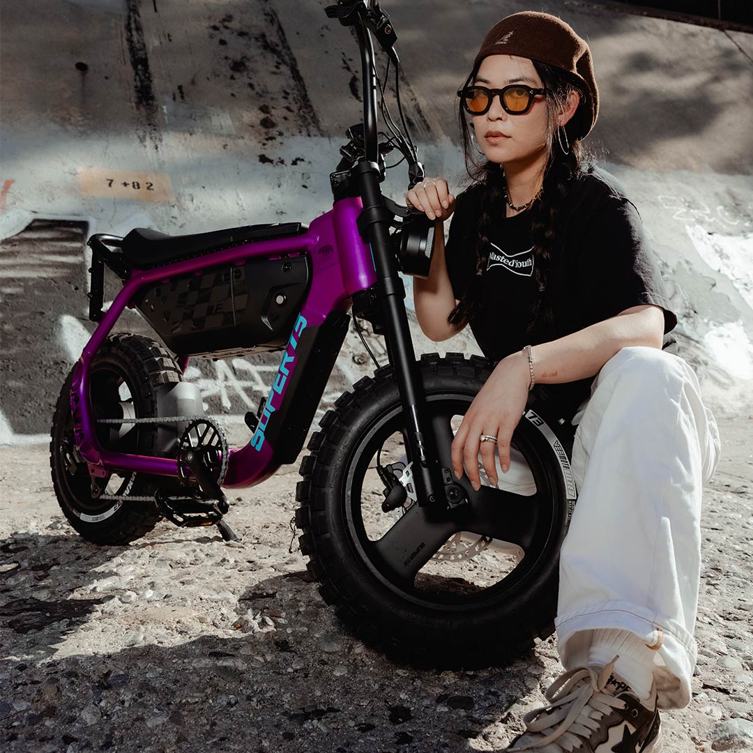 A woman wearing white jeans and a black t-shirt kneels over the front wheel of a SUPER73-MZFT ebike parked in a concrete riverbed.