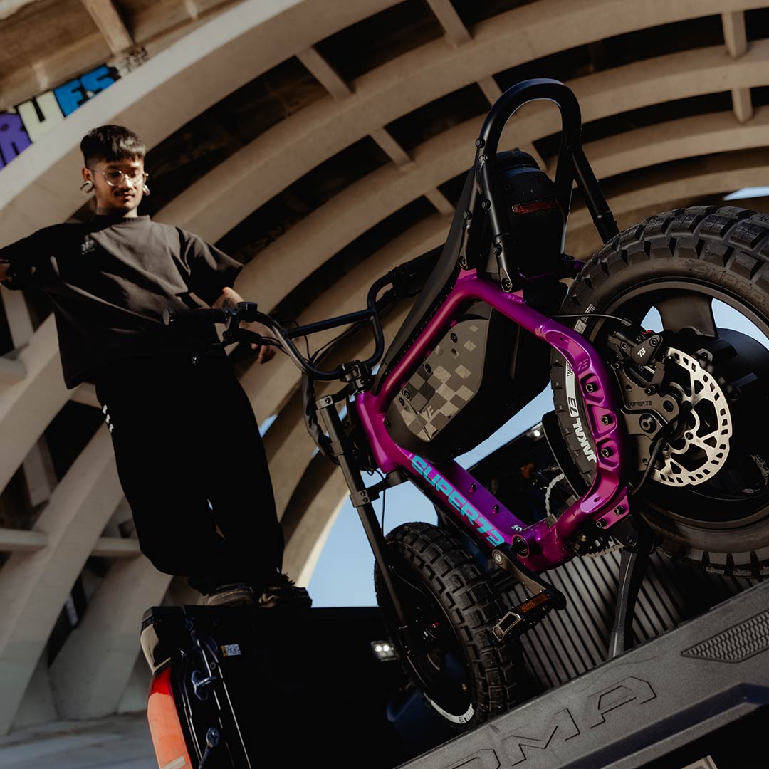 A man with tattoos jumps down from the bed of a Toyota Tacoma truck. A SUPER73-MZFT ebike in Cullenberry is loaded into the bed of the truck behind the man.