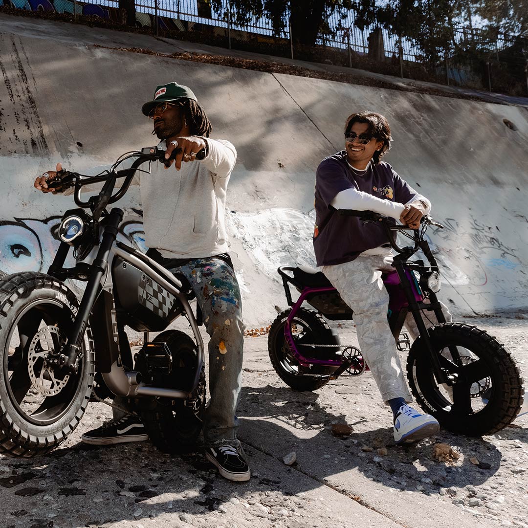 A man sits on the right wearing light colored pants and a white long sleeved shirt underneath a dark t-shirt. Another man sits on the left wearing paint splattered jeans and a gray sweatshirt. Both are sitting on SUPER73-MZFT ebikes parked on a concrete riverbed.