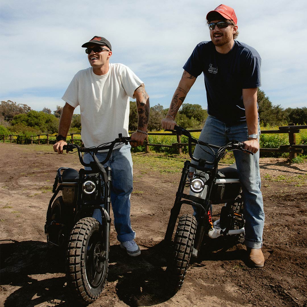 Two men, both in jeans and t-shirts, standing on a dirt path with green fields and a wooden fence in the background. The man on the left is standing next to a SUPER73-MZFT SE ebike in Blackout and the man on the right is straddling a SUPER73-MZFT SE ebike in Baja.