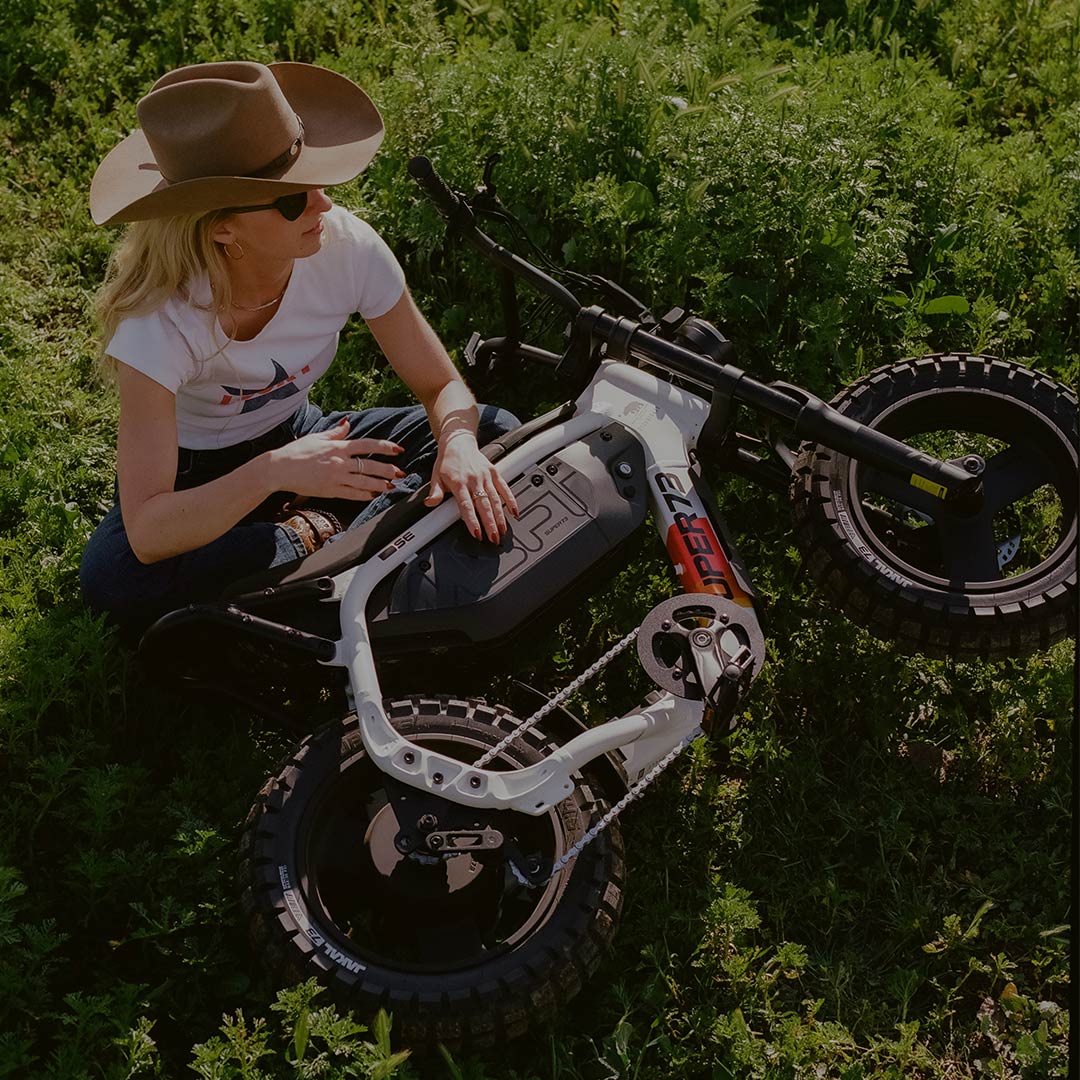 A woman wearing dark jeans, a white t-shirt, and a brown cowboy hat is sitting cross-legged on the ground in a green field. A SUPER73-MZFT SE ebike is laid on its side in front of her and she has one hand on the frame of the bike.