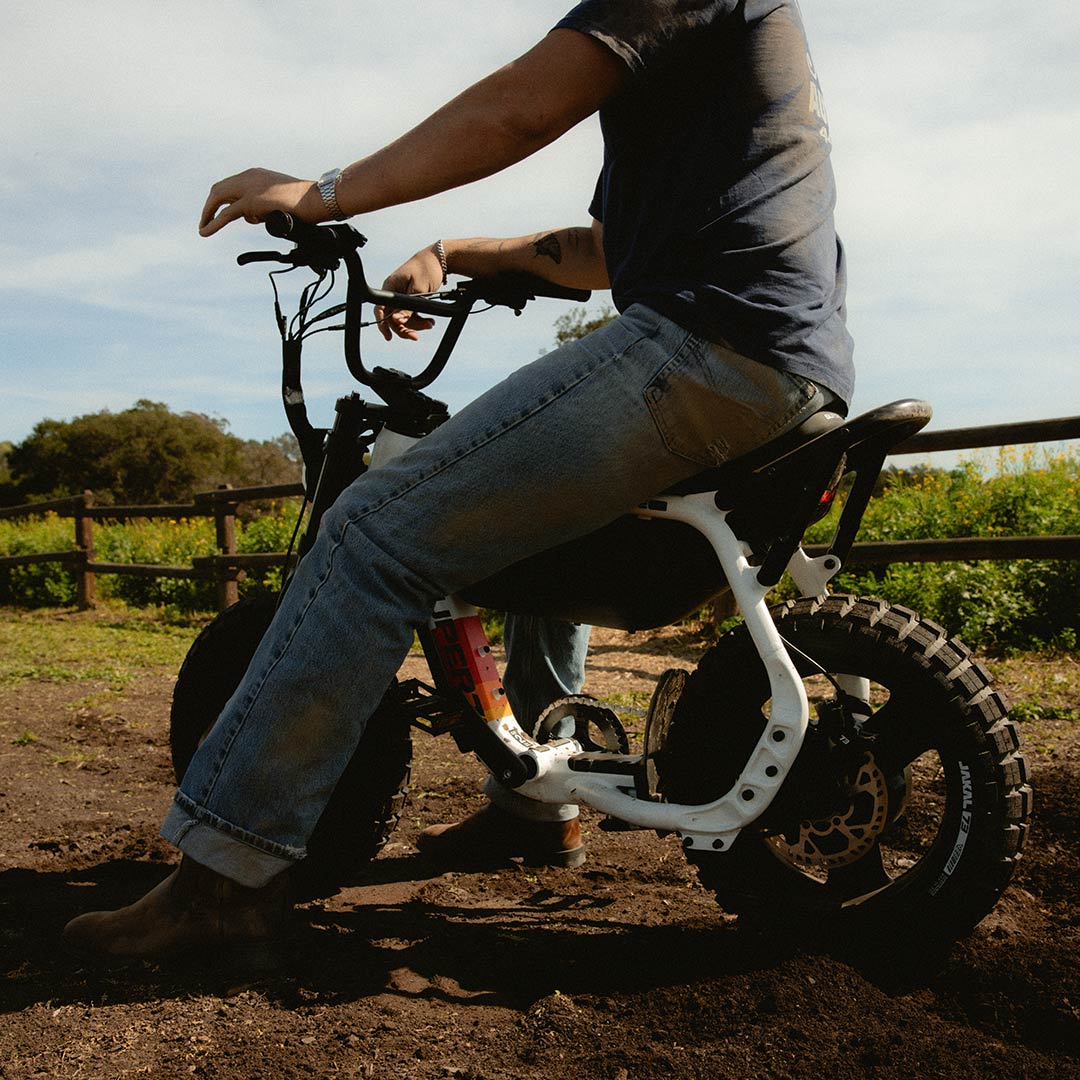 A man wearing jeans and a t-shirt, who is slightly cropped out at the top, is sitting on a SUPER73-MZFT SE ebike on a dirt path. A green field and wooden fence are visible in the background.