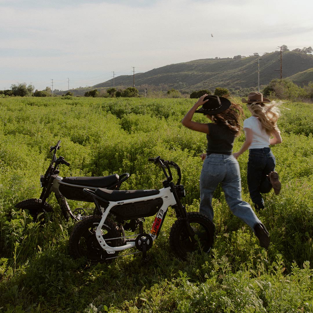 Two women, both in jeans and t-shirts, are standing in a large green field with their backs to the camera. Two SUPER-73 MZFT SE ebikes are parked in the field, just to the left of the women.
