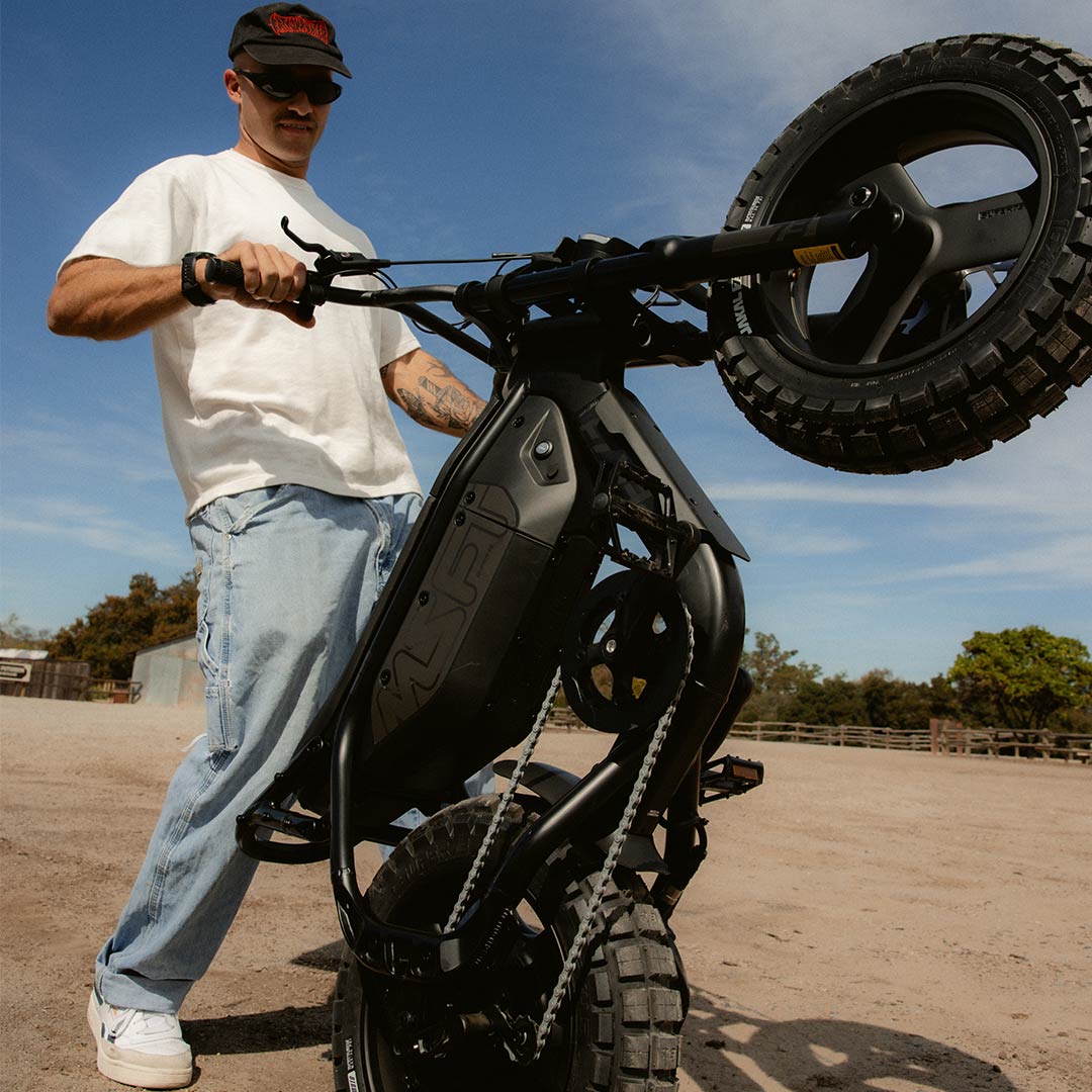 A man wearing jeans and a white t-shirt standing on a dirt path and holding a SUPER73-MZFT SE in a wheelie position.