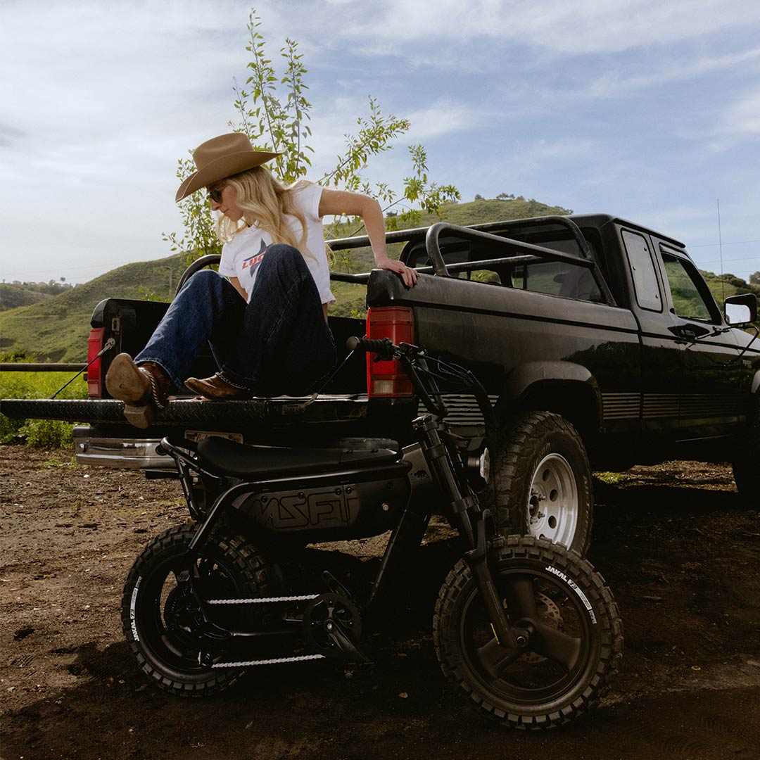 A woman wearing jeans, a white t-shirt, and a brown cowboy hat is sitting on the tailgate of a pickup truck parked in front of a green hill. A SUPER73-MZFT is parked on the dirt in front of her.