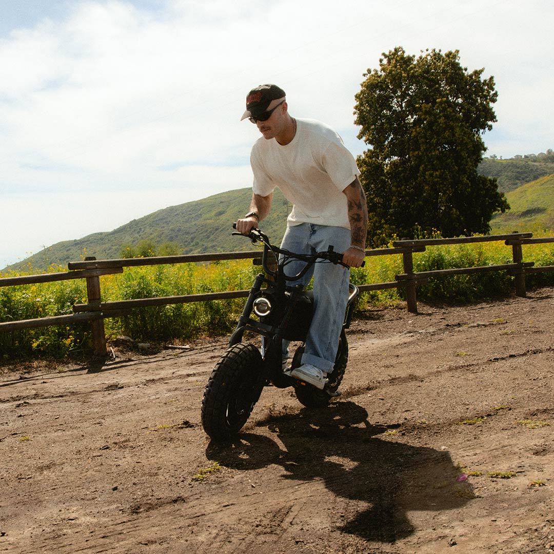A man in jeans and a white t-shirt riding a SUPER73-MZFT SE on a dirt path with green hills in the background.