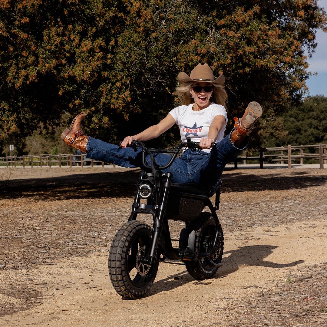 A woman in jeans, a white t-shirt, and a cowboy hat is riding a SUPER73-MZFT SE along a dirt path with a large tree and wooden fence in the background. She is kicking both of her feet up and smiling.