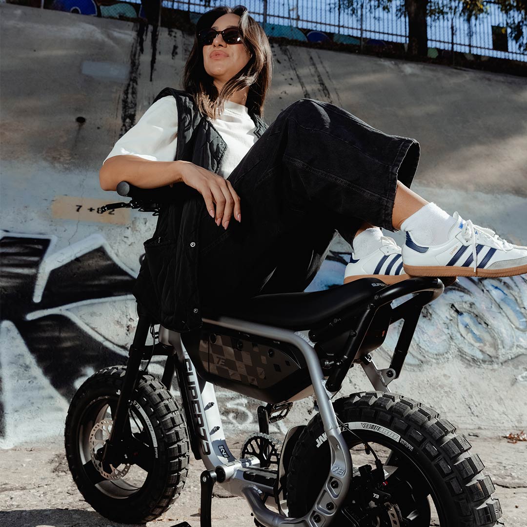 A woman in black jeans and a white t-shirt sits backwards on top of a SUPER73-MZFT ebike. The bike is parked in a concrete riverbed and graffiti is visible behind the woman.