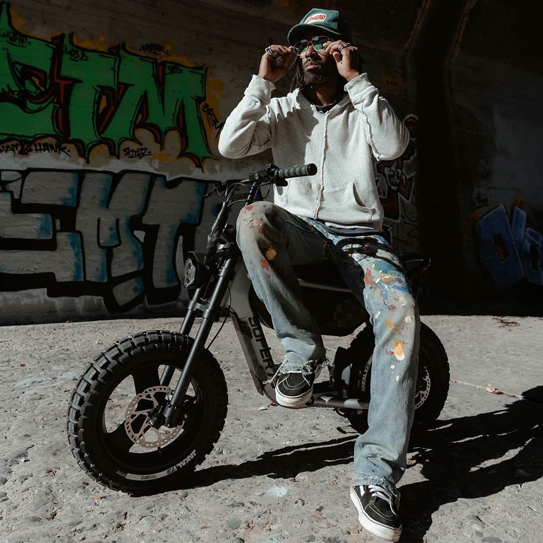 A man wearing a gray sweatshirt and paint splattered jeans is sitting sideways on the SUPER73-MZFT in Titanium Ore. The bike is parked in a concrete riverbed with graffiti in the background.