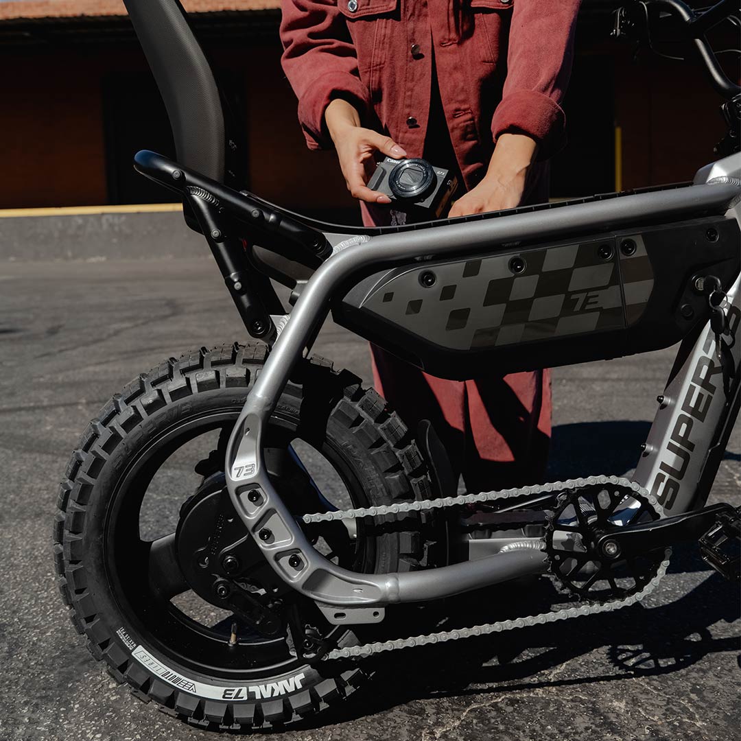 A SUPER73-MZFT ebike is parked on asphalt in the foreground while a person wearing a red jean jacket and matching pants stands behind the bike holding a digital camera.