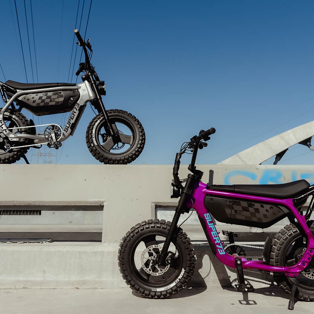 On the left, a SUPER73-MZFT ebike in Titanium Ore is parked atop a concrete wall with power lines stretching out in the background. On the right, a SUPER73-MZFT ebike in Cullenberry is parked at ground level in front of the wall.